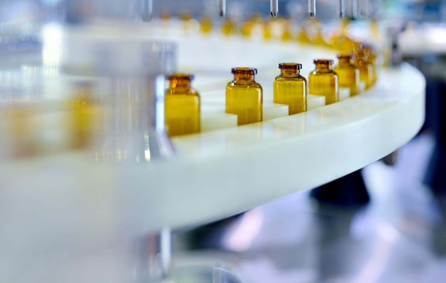Empty amber glass vials move along a white industrial conveyor belt in a pharmaceutical manufacturing facility production line.
