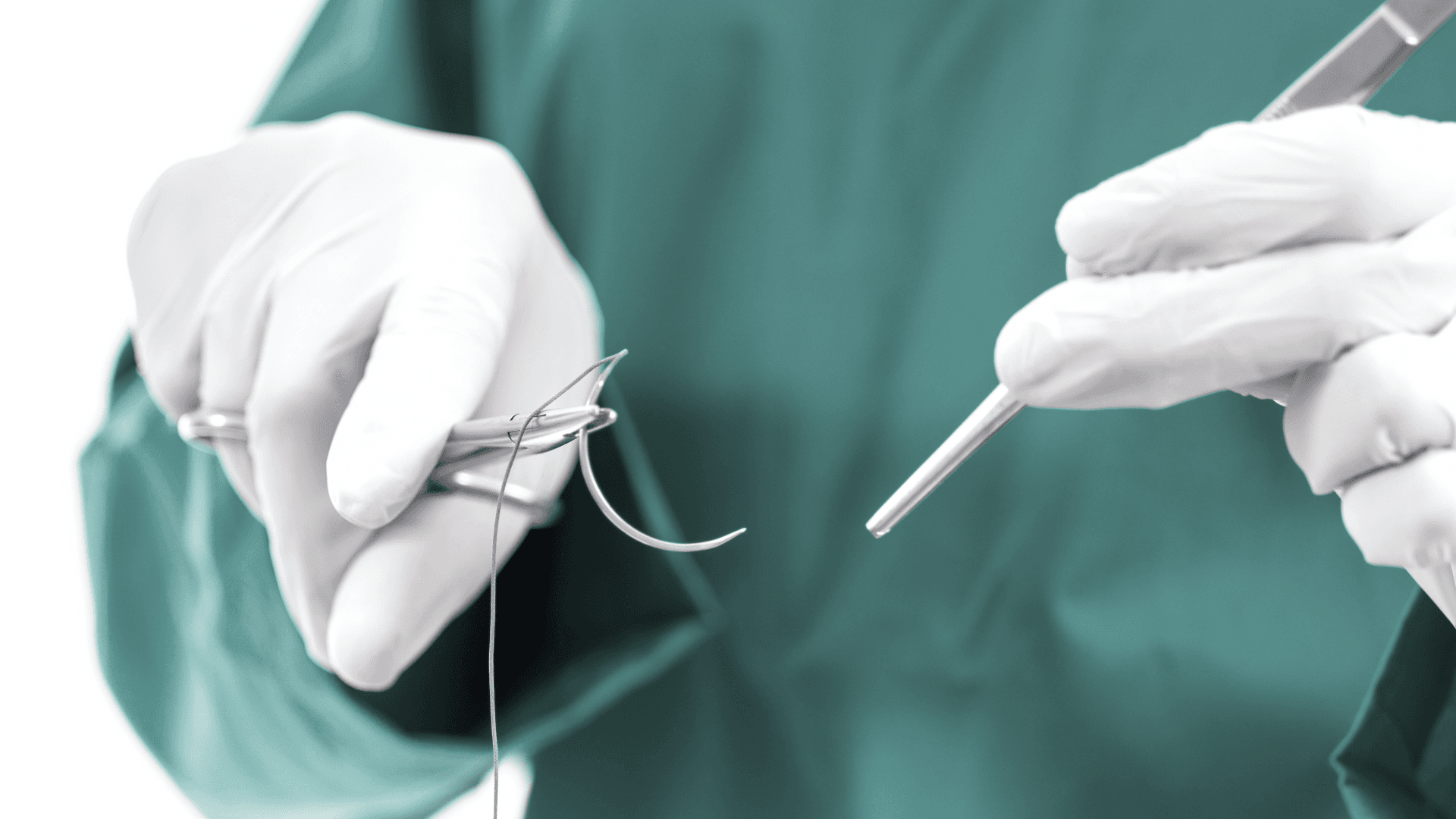Surgeon in green scrubs and white gloves holds a needle driver with a curved suture needle and thread during a medical procedure.