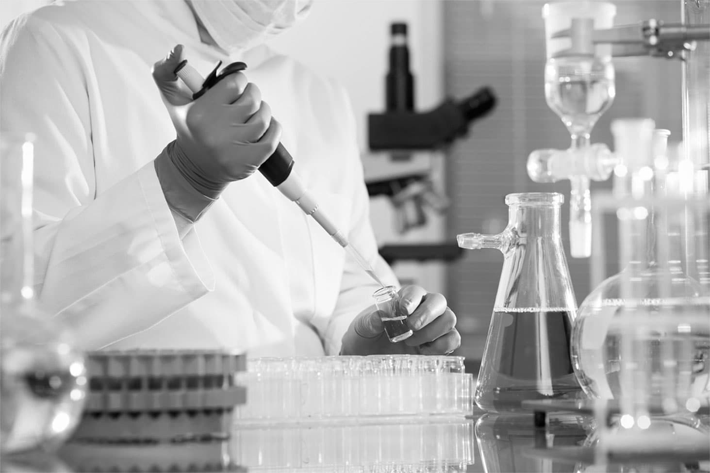 A laboratory technician in a white coat uses a pipette to transfer liquid into a small vial amidst various glass laboratory equipment.