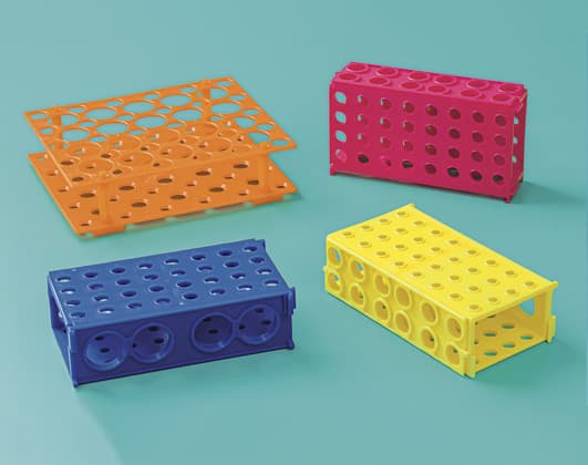 Four colorful plastic laboratory test tube racks in orange, pink, blue, and yellow arranged on a solid light blue background.