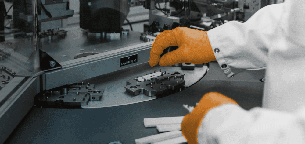A technician wearing orange gloves operates an automated manufacturing machine in a clean room environment, handling small white medical test components.