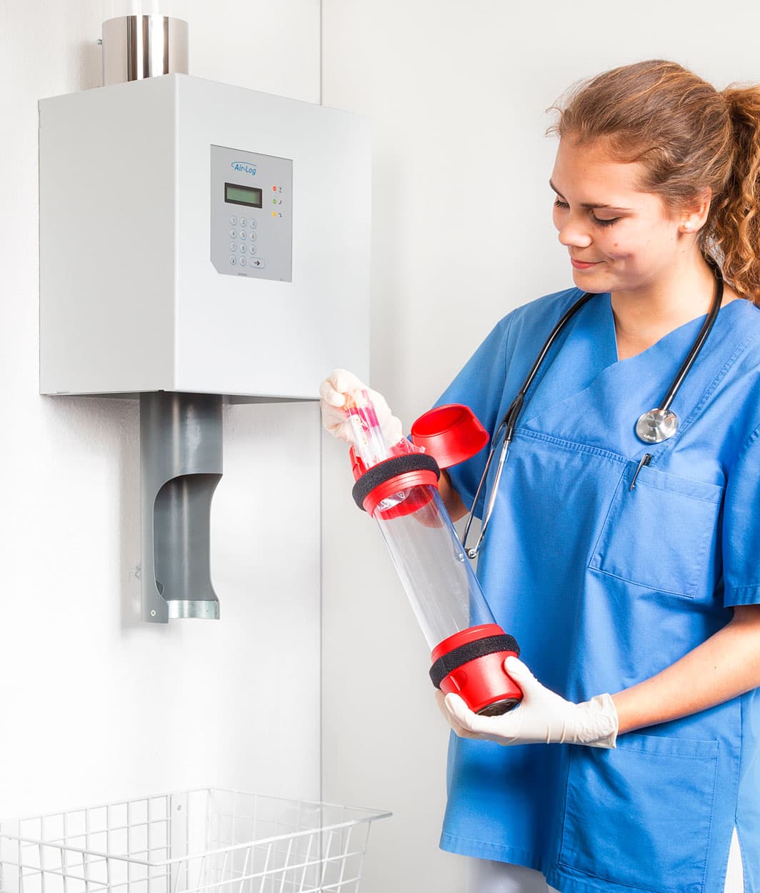 A healthcare professional in blue scrubs inserts a pneumatic tube carrier containing medical samples into an Air-Log wall-mounted station.