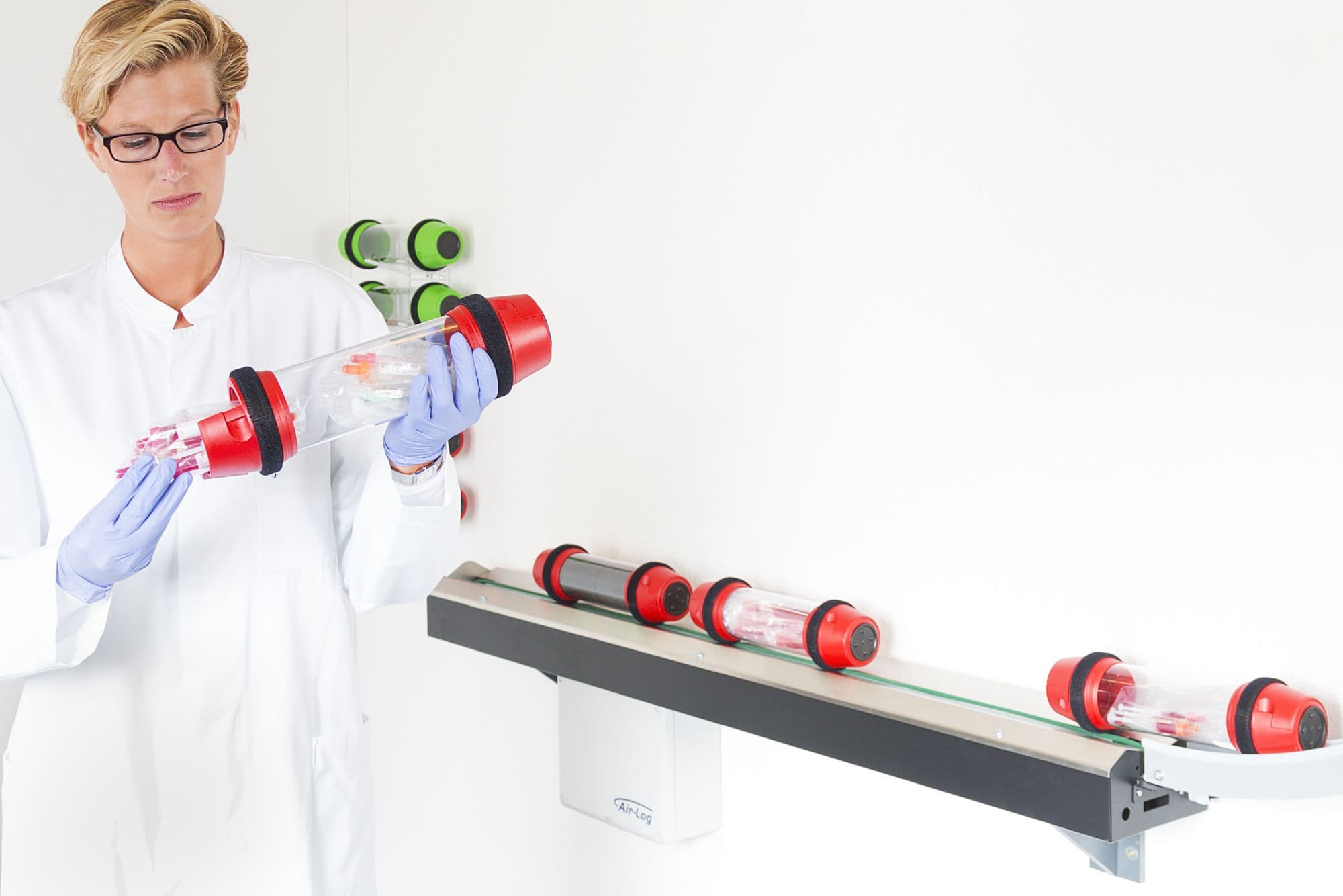 A laboratory technician in a white coat and blue gloves handles a pneumatic tube carrier near a wall-mounted transport track system.