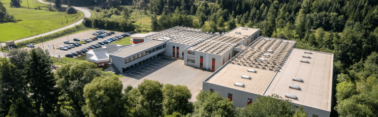 Aerial view of a large industrial facility with solar panels on the roof, parking lot, and surrounding green forest landscape.