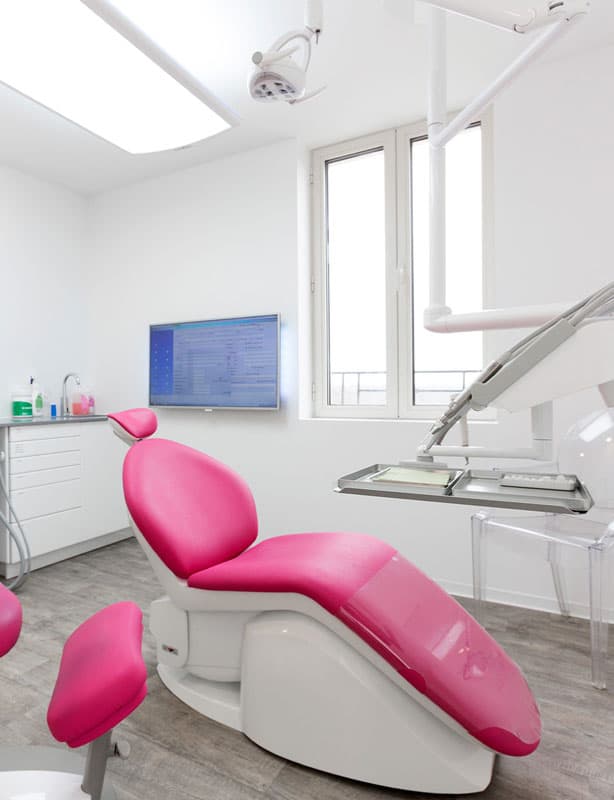 A modern dental operatory featuring a bright pink patient chair, overhead lighting, and a wall-mounted digital display screen for patient records.