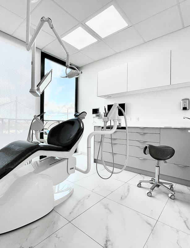 Modern dental operatory featuring a black patient chair, overhead lighting, integrated dental delivery unit, and ergonomic stool in a bright clinical setting.