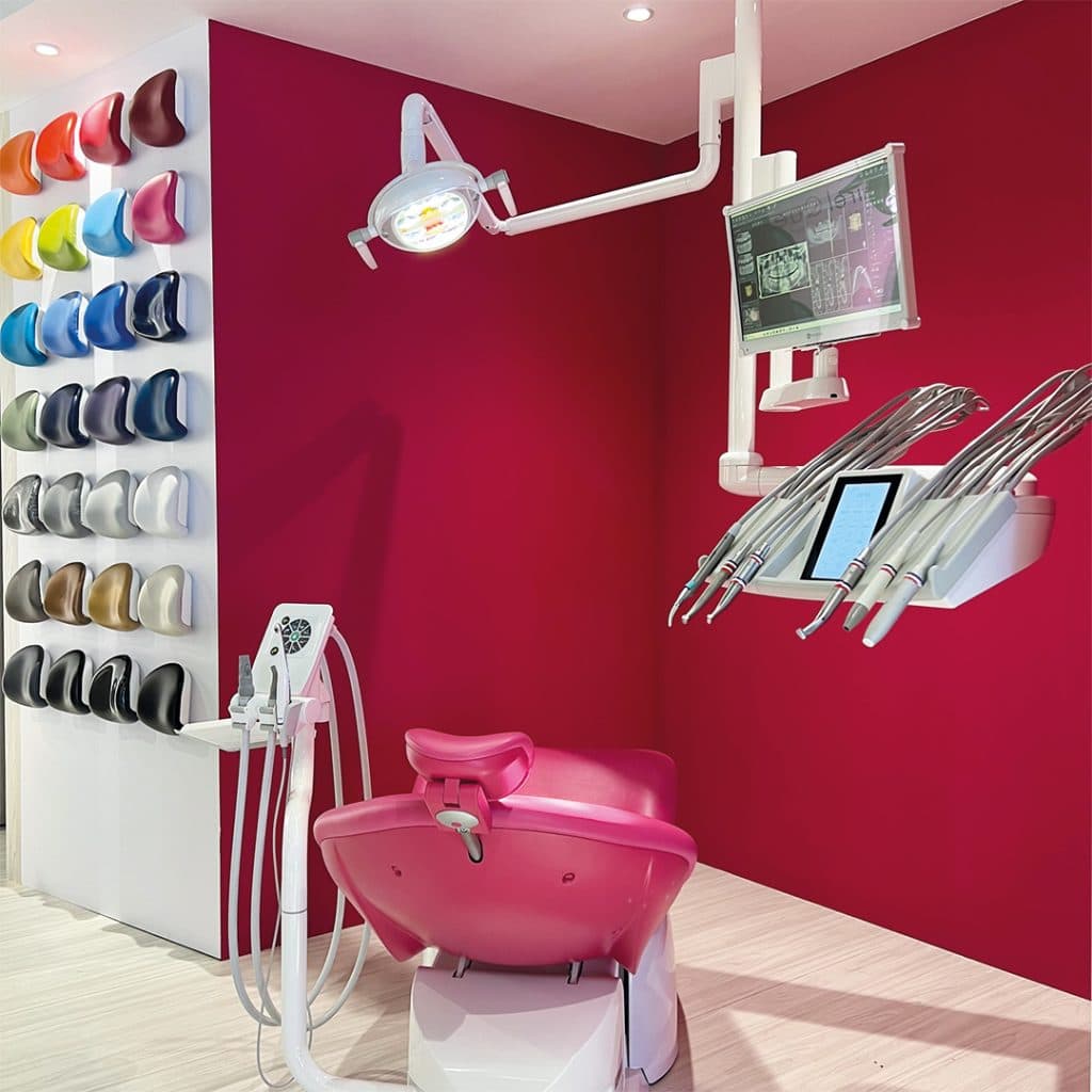A dental operatory setup featuring a pink patient chair, overhead light, digital display, and a wall-mounted display of various colored headrest upholstery options.