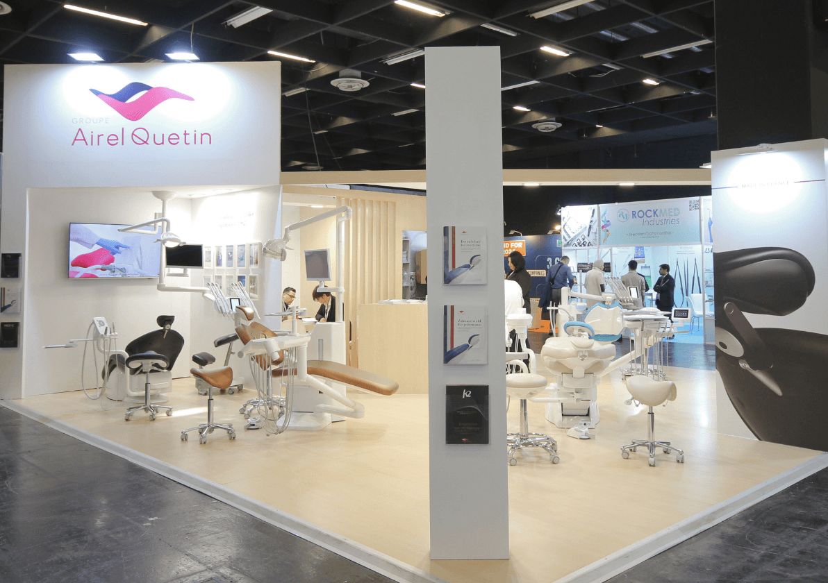 A trade show booth featuring multiple dental chairs, stools, and equipment displays under bright lighting with company branding visible overhead.
