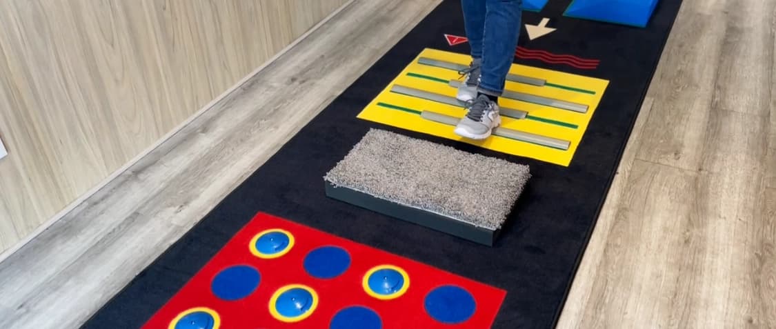 A person in jeans and sneakers walks across a physical therapy gait training mat featuring various textured surfaces and colored tactile markers.