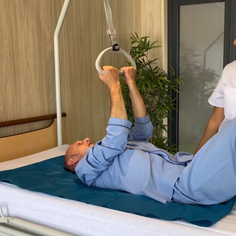 A patient in a hospital bed uses a white overhead trapeze bar to assist with mobility while a caregiver stands nearby.
