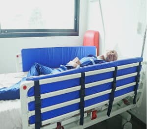 A patient lies in a hospital bed equipped with blue protective side rail padding secured by black adjustable straps.