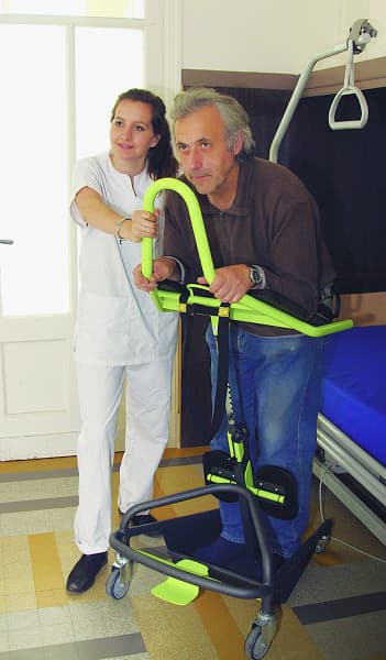 A caregiver assists a patient using a bright green mobile standing aid device in a clinical room setting.