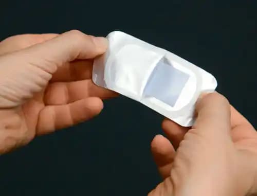 Hands holding a single small, rectangular medical patch in its silver foil packaging against a dark background.