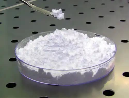 A metal spatula lifts a sample of white powder from a petri dish resting on a perforated stainless steel laboratory workbench surface.
