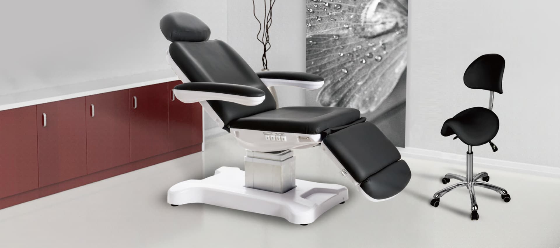 A black and white medical treatment chair and matching stool positioned in a studio setting with a decorative wall print.