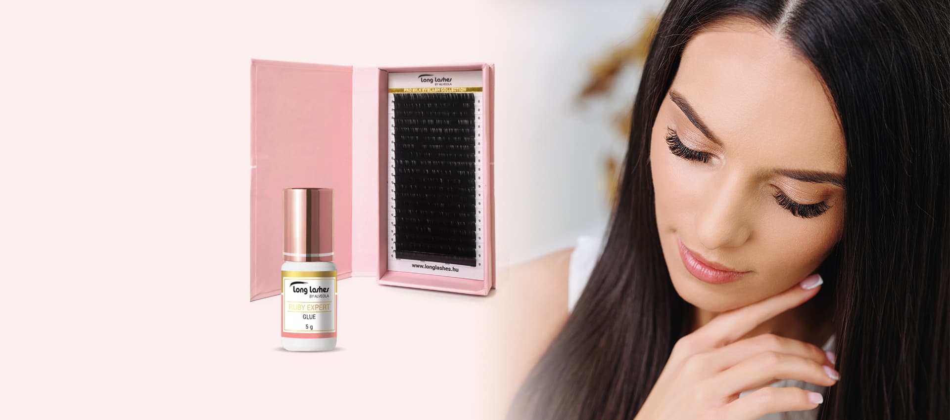 A woman with long eyelash extensions poses next to a pink box containing a tray of lashes and a small glue bottle.