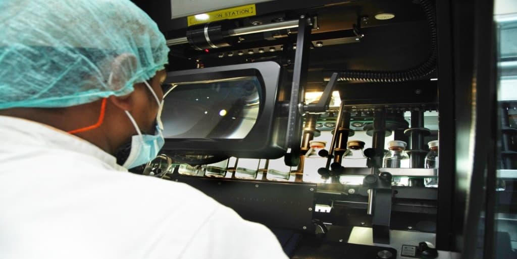 Technician in sterile gear monitoring an automated pharmaceutical filling line for injectable solutions in a clean room.