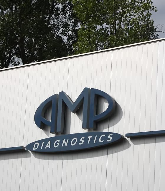 Exterior building wall featuring the blue three-dimensional AMP Diagnostics company logo mounted on white vertical siding panels.