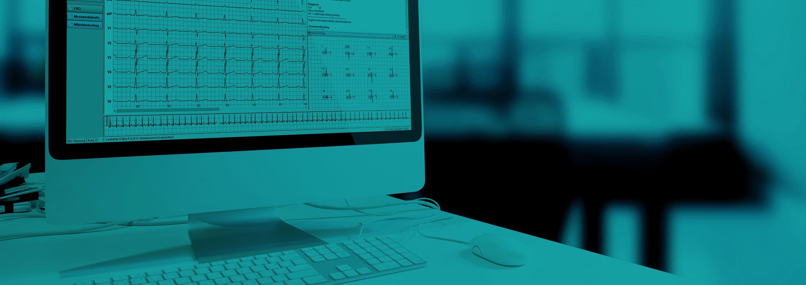 Desktop computer monitor displaying ECG rhythm traces and diagnostic data grids on a clinical office desk with keyboard and mouse.