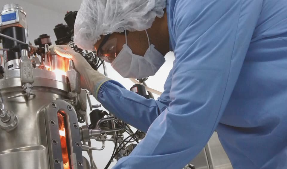 Scientist in clean room attire monitoring a stainless steel bioreactor during the manufacturing process.