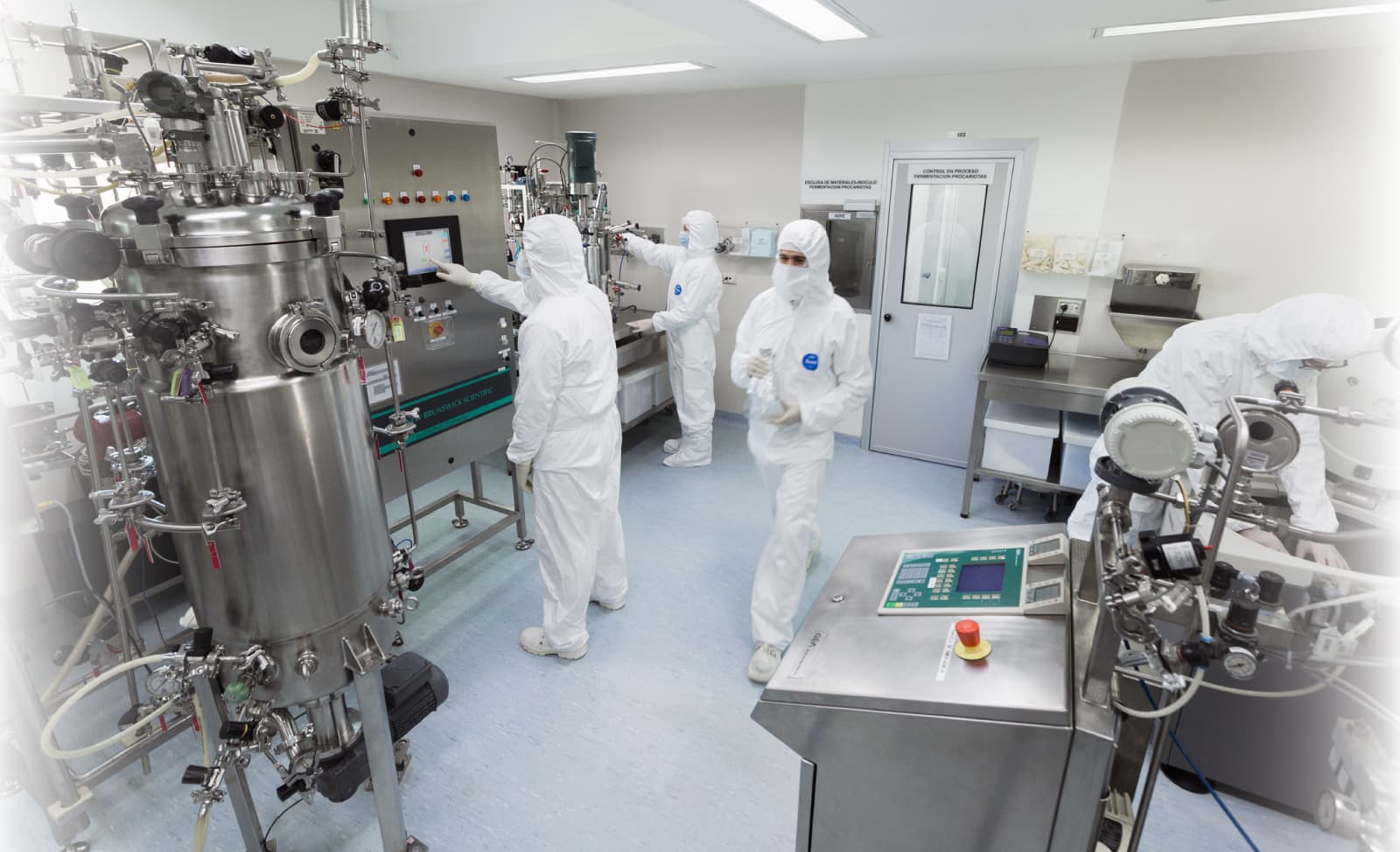 Technicians in white cleanroom suits operate stainless steel bioreactors and control panels within a sterile pharmaceutical manufacturing laboratory environment.