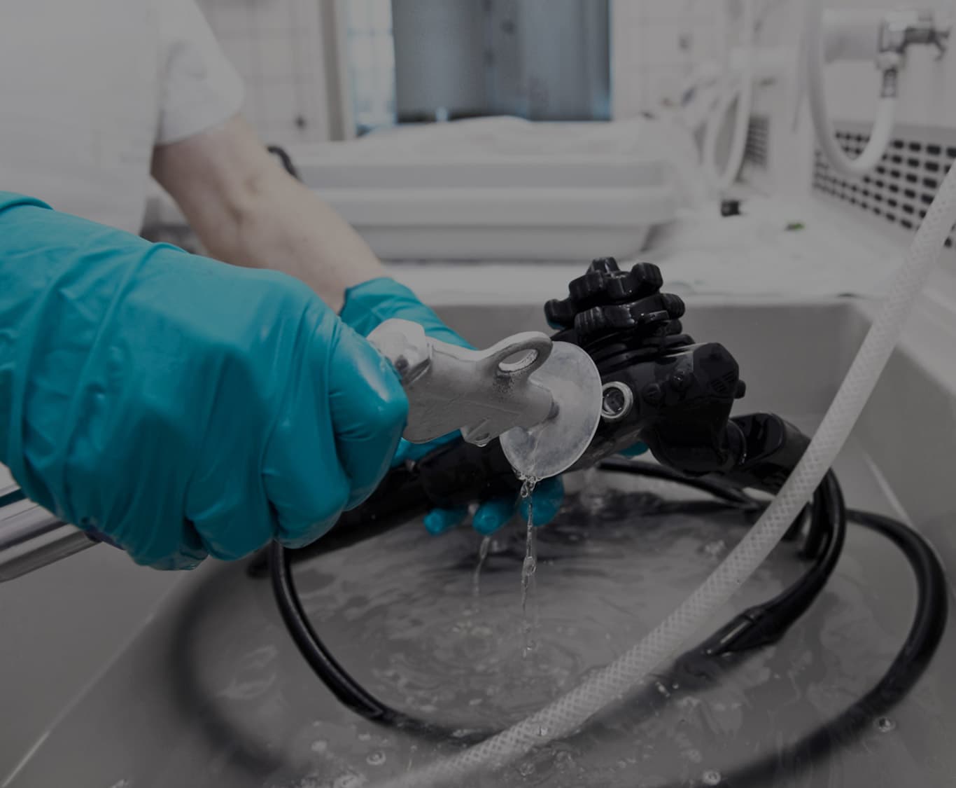 A healthcare worker wearing blue nitrile gloves rinses a flexible endoscope in a stainless steel sink within a clinical reprocessing area.