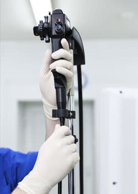 A healthcare professional wearing white latex gloves handles a black medical endoscope device mounted on a metal stand in a clinical setting.