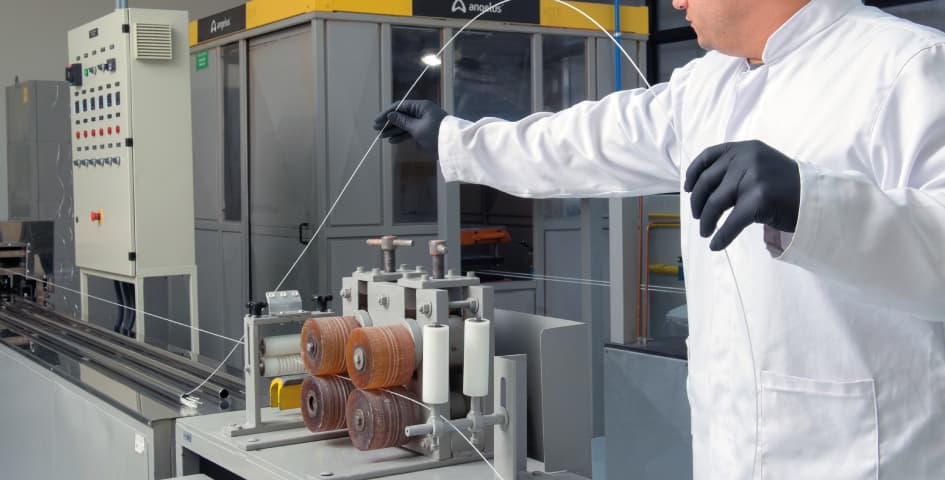 A technician in a white lab coat and black gloves monitors a wire-drawing machine on a manufacturing production floor.