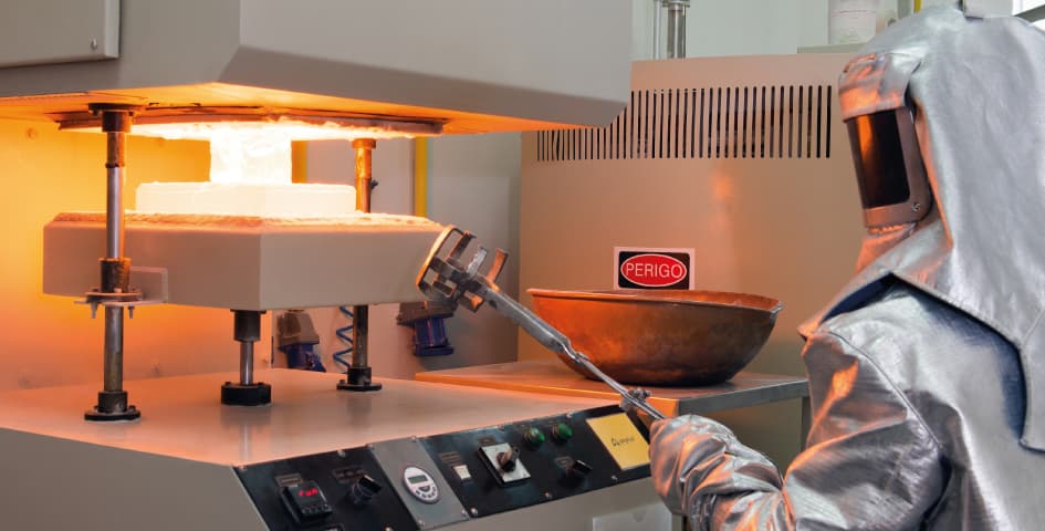 A worker in protective silver gear operates a high-temperature industrial press machine within a manufacturing facility, handling glowing materials with tongs.