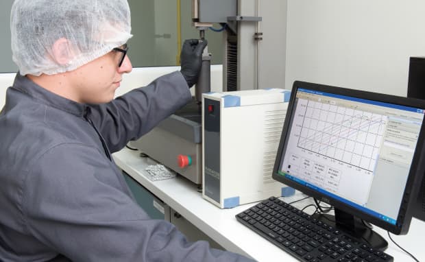 A technician in a hairnet and lab coat operates a testing machine while monitoring real-time data on a computer screen in a laboratory.