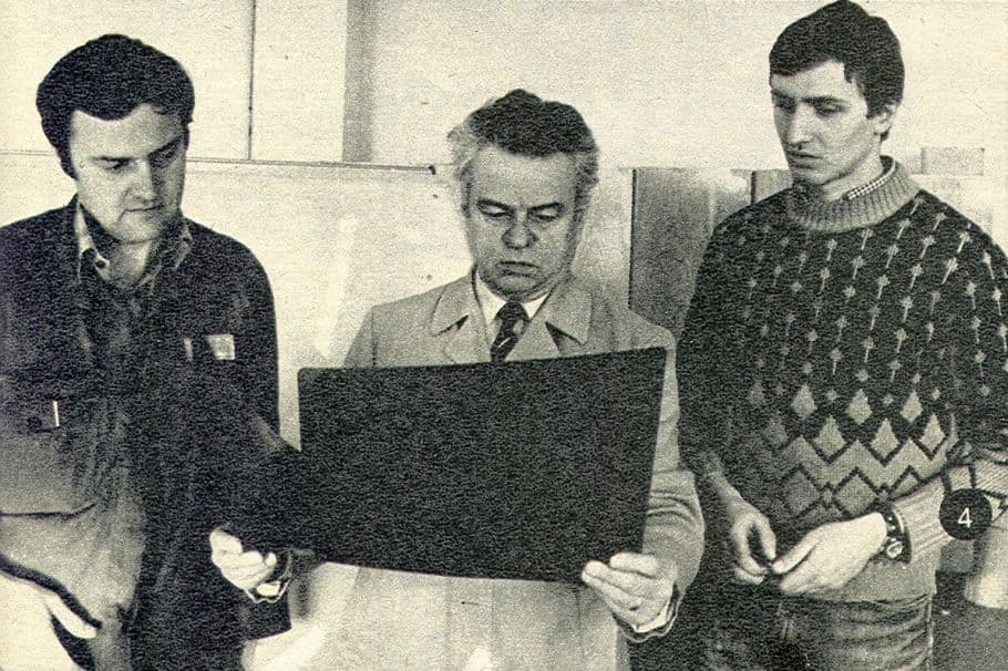 Three men in 1980s attire examine a large dark document or film in an office or workshop setting.