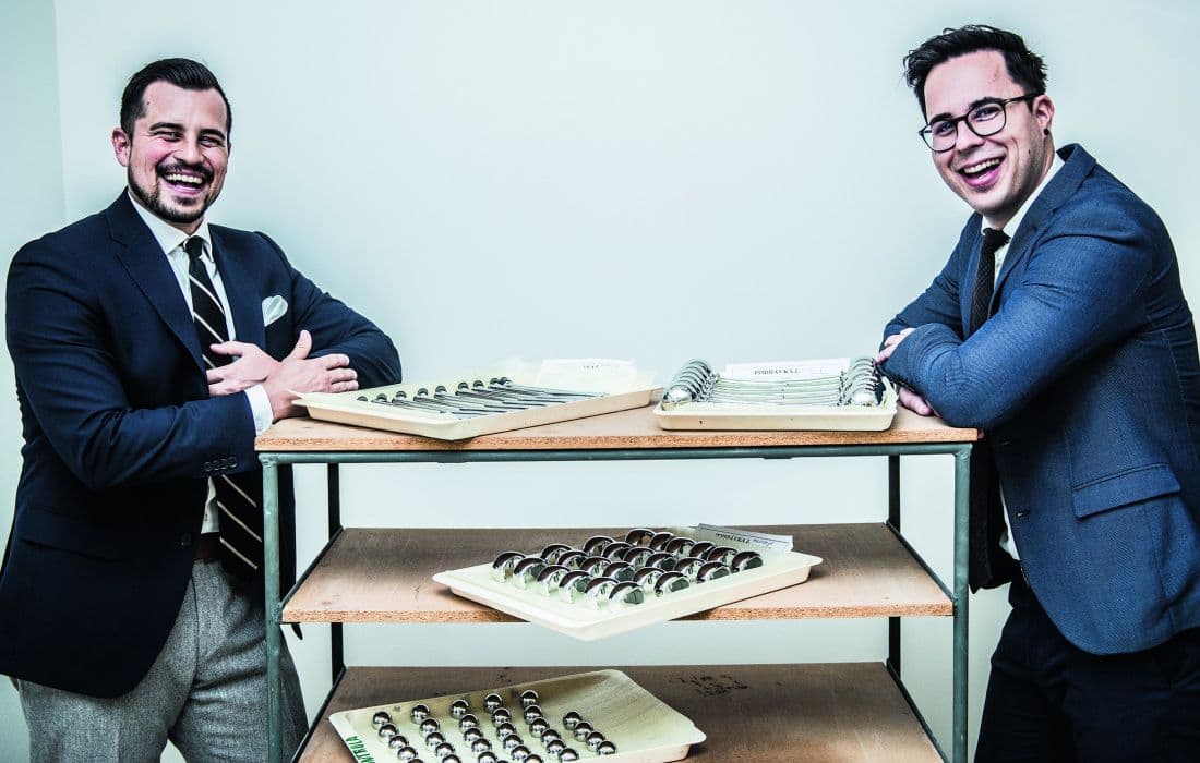 Two smiling men in business attire stand on either side of a metal shelving unit displaying multiple trays of orthopedic medical components.
