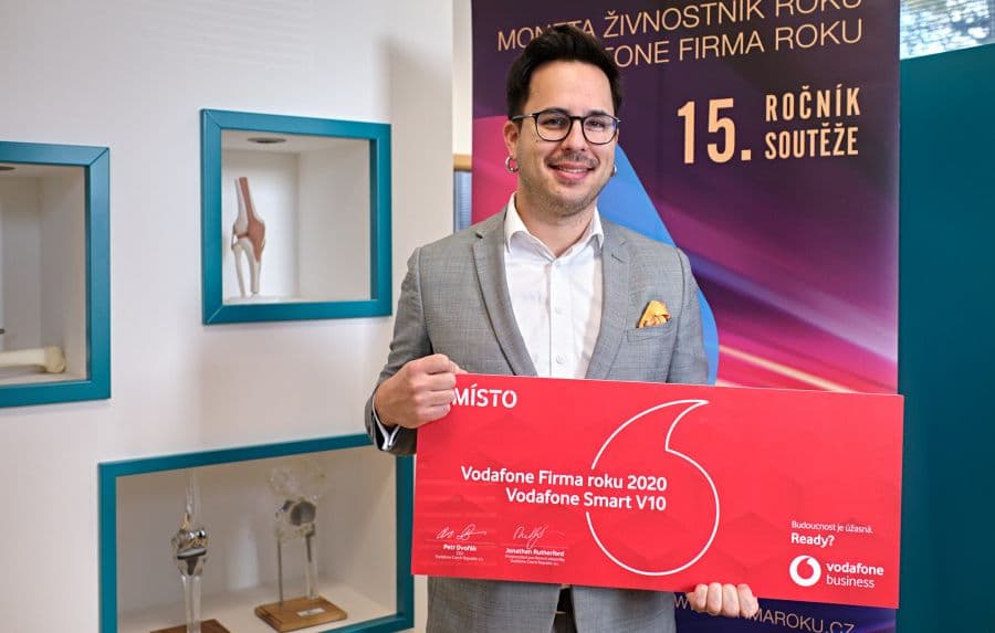 A man in a grey suit holds a red award plaque for a business competition, standing before company display shelves.