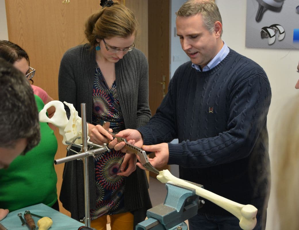 A group of people in a workshop setting examining a metal orthopedic implant component against a plastic bone model.