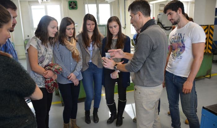 A group of young adults observes a male instructor demonstrating a small medical component inside a bright industrial manufacturing facility.