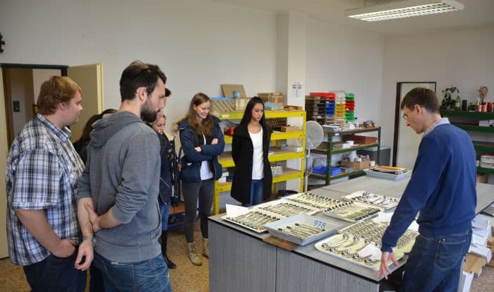 A group of young adults observes a man demonstrating various orthopedic implant components arranged on trays in a workshop or training room.