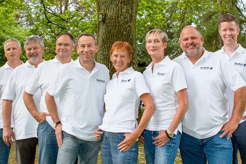 A group of eight staff members wearing white branded polo shirts stand together outdoors in front of a large tree.