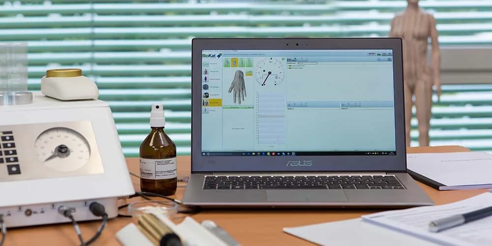 A laptop displays the BioKat software interface featuring a hand model and diagnostic gauges, positioned on a desk with medical equipment.