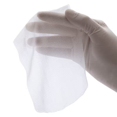 A gloved hand holds a single sheet of white, semi-transparent, woven hemostatic gauze against a plain white background.