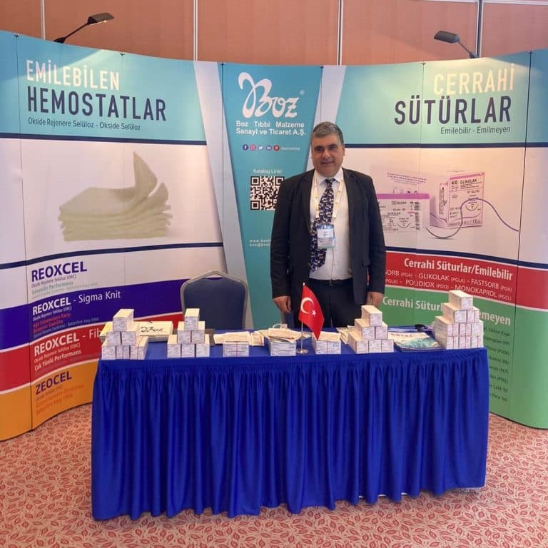 A company representative stands behind a blue-clothed table displaying multiple medical product boxes at a surgical congress trade show booth.