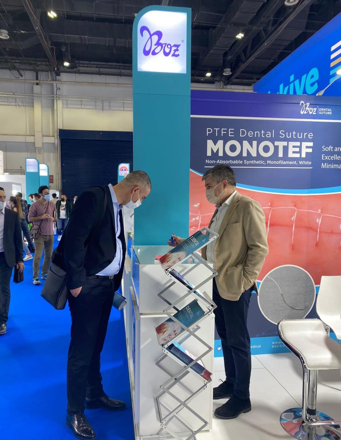 Two men in business attire discuss products at a branded trade show booth featuring dental suture signage and marketing literature.