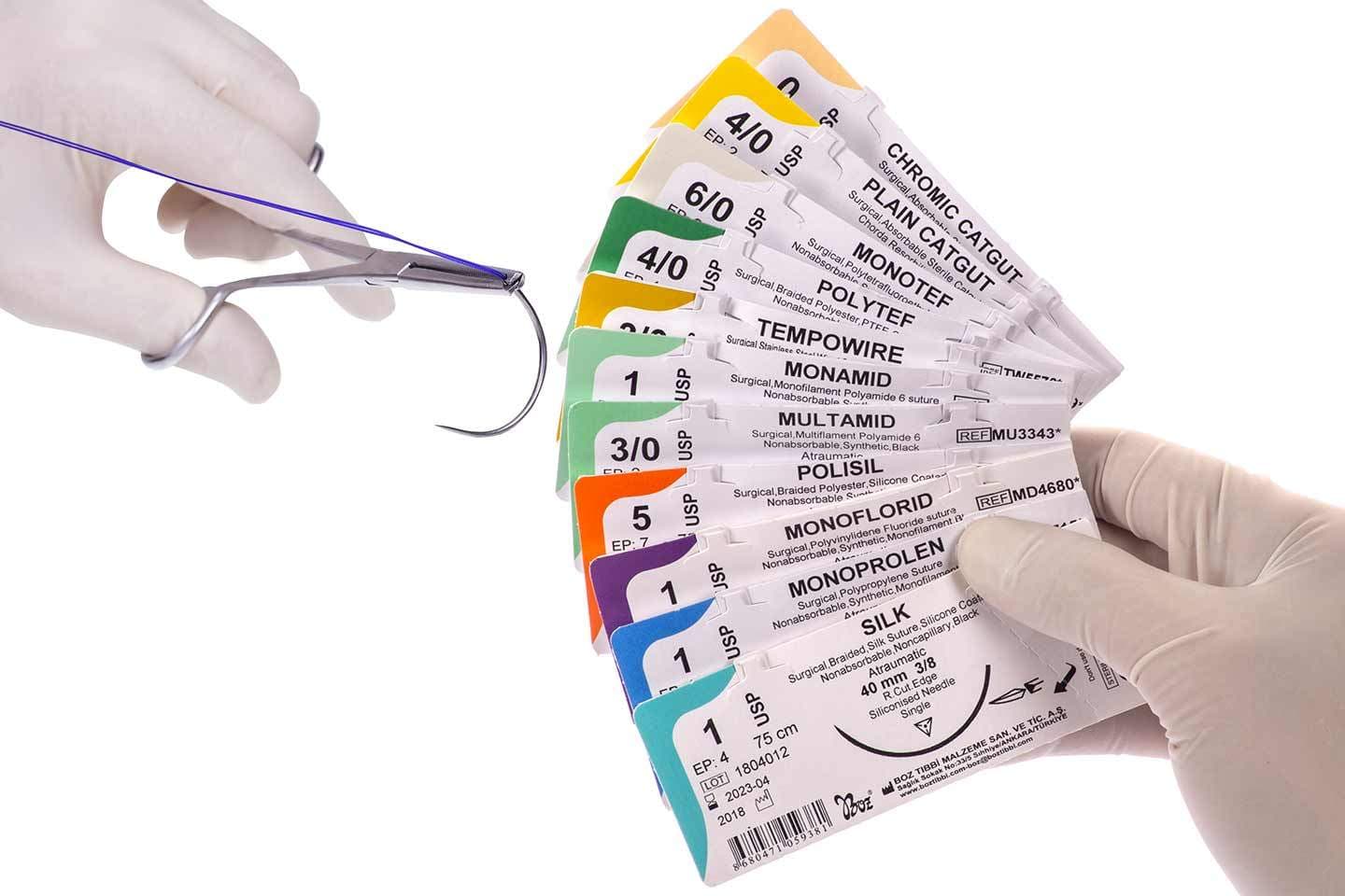 A gloved hand fans out a variety of color-coded surgical suture packets while another hand holds a needle with blue thread.