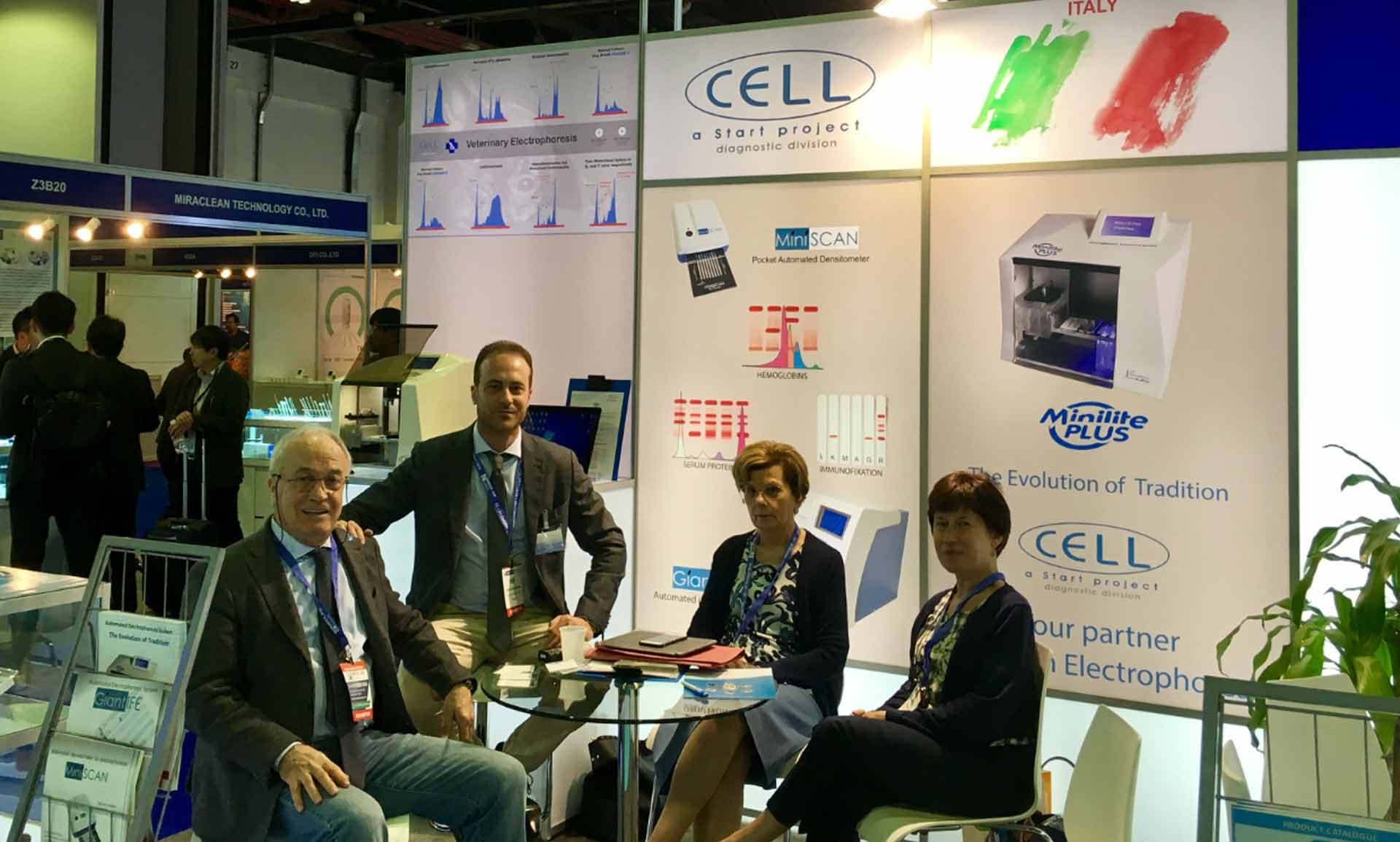 Four professionals sit at a table in a trade show booth featuring company branding, product posters, and diagnostic equipment displays.