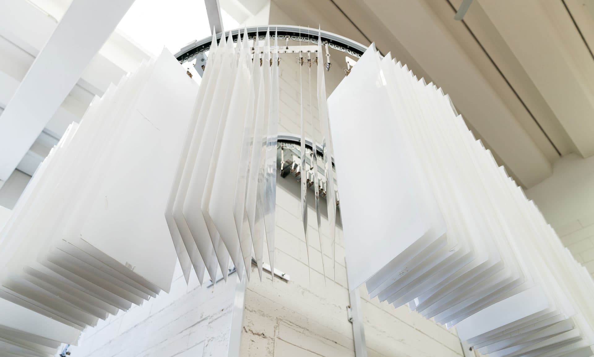 A circular industrial conveyor system suspends multiple white rectangular panels in a bright, clean manufacturing or laboratory facility environment.