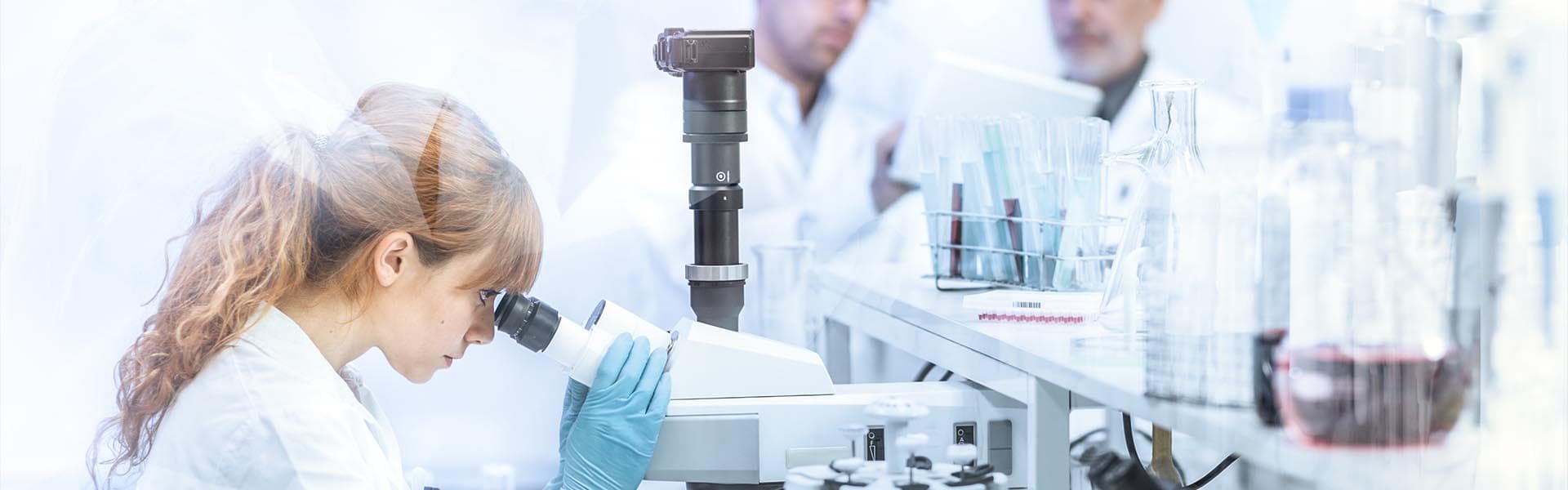 A researcher in a lab coat uses a microscope while colleagues work in the blurred background, featuring laboratory glassware and equipment.