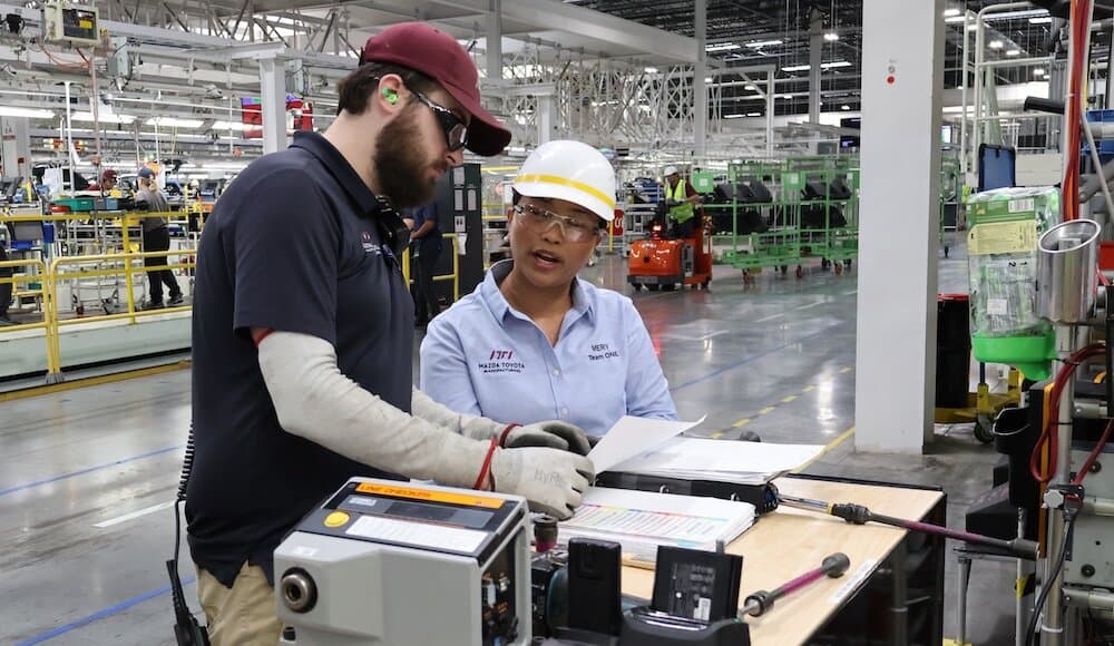 Two employees in safety gear reviewing technical documentation at a manufacturing workstation.