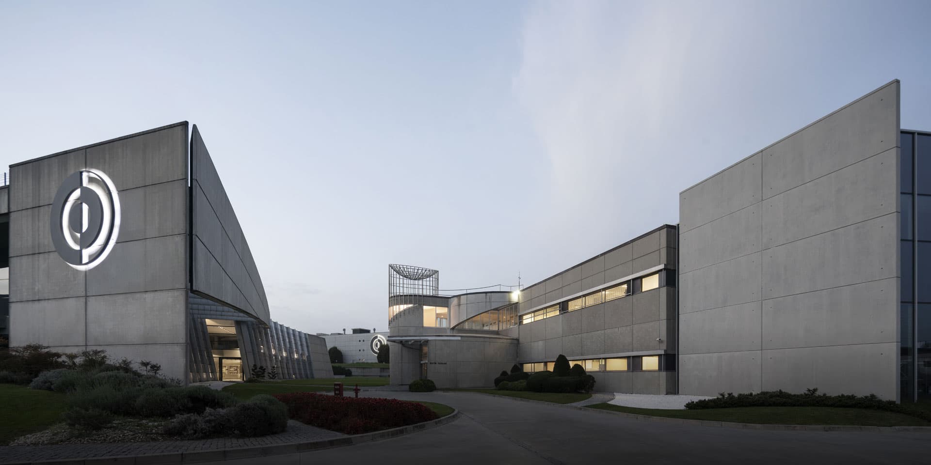 Modern concrete pharmaceutical manufacturing facility exterior featuring angular architecture, landscaped grounds, and a large circular company logo on the facade.