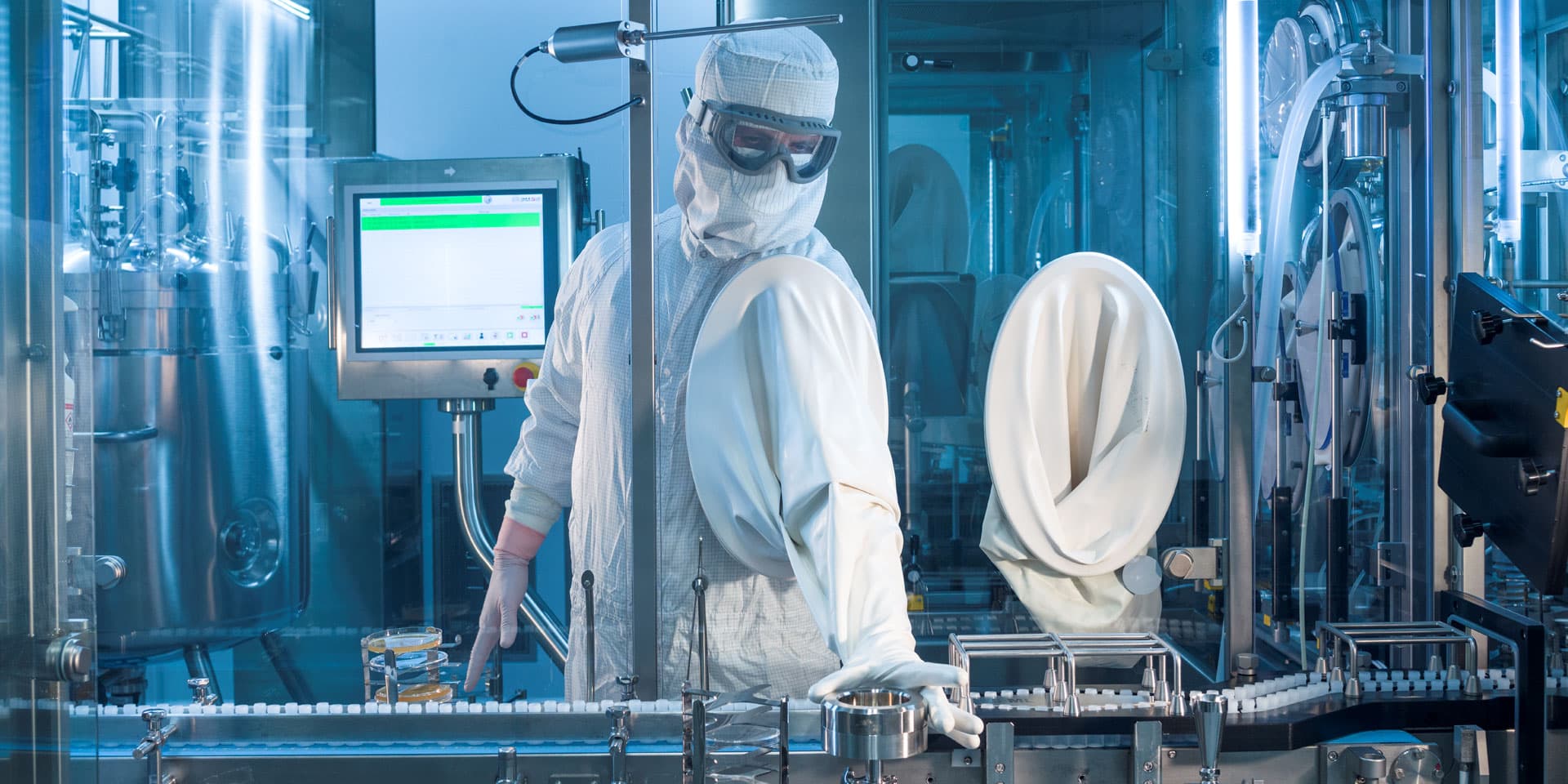 A technician in full clean room protective gear operates a pharmaceutical production line with stainless steel machinery and integrated control screens.